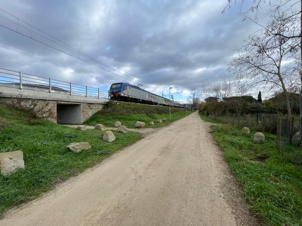 Tratto pianeggiante della ciclovia parallelo alla ferrovia, a sinistra, e recinzione a destra. Rocce a terra ai bordi del percorso.